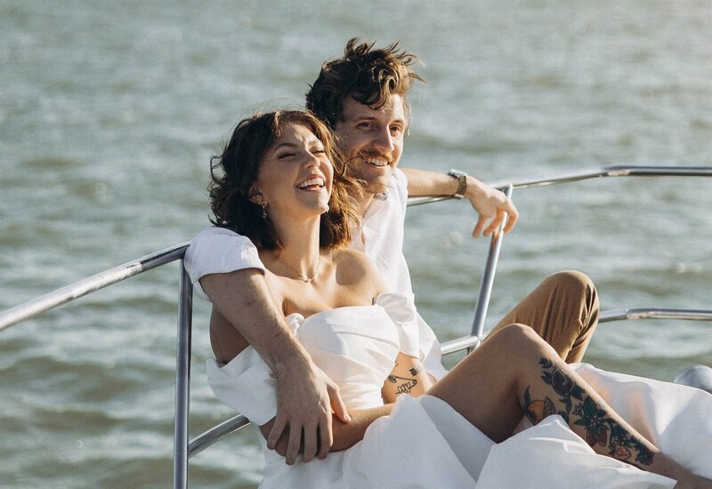 Groom throws his arm around bride's shoulders as they laugh on yacht.