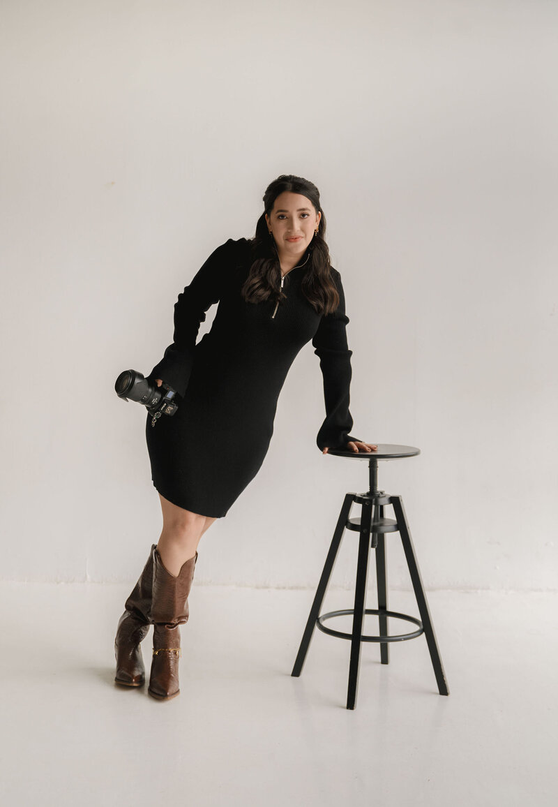 Houston photographer in a black dress and brown boots leans on a stool, holding a camera. The minimalist, bright setting conveys a poised, professional tone.