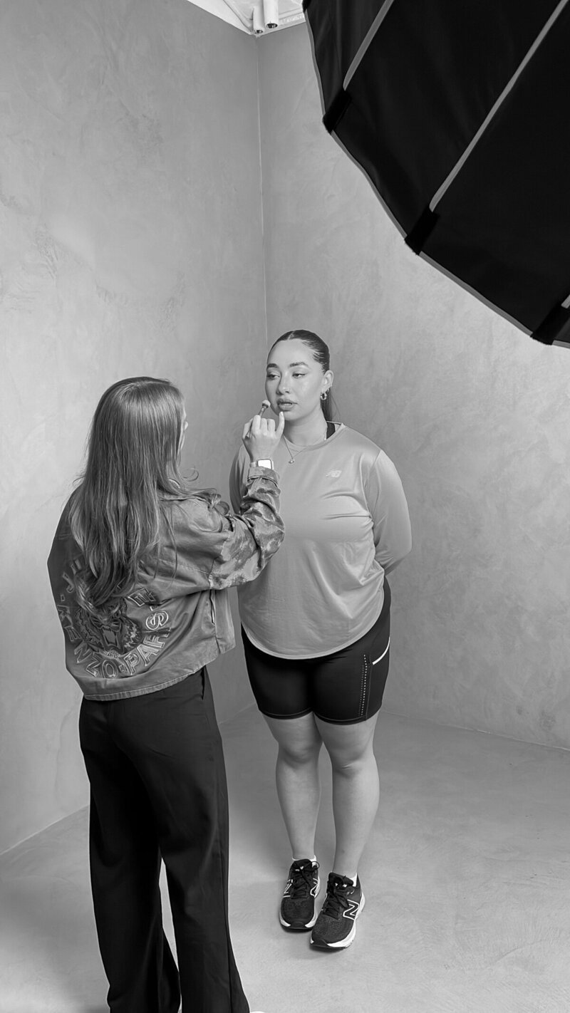 Abby Kaye applying makeup behind the scenes of a New Balance athletic photoshoot, with a model dressed in workout attire.