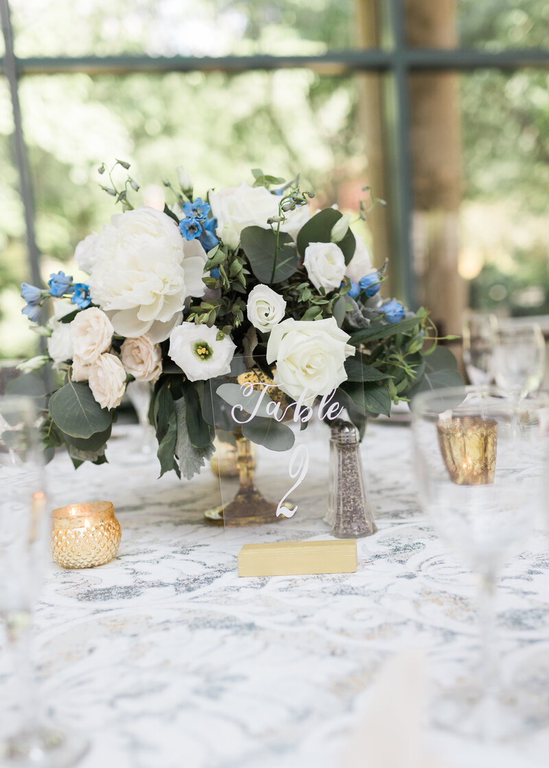 textured dusty blue and gold flecked floral linen with white and blush florals in a gold compote behind an acrylic table number #2 surrounded by gold votive candles