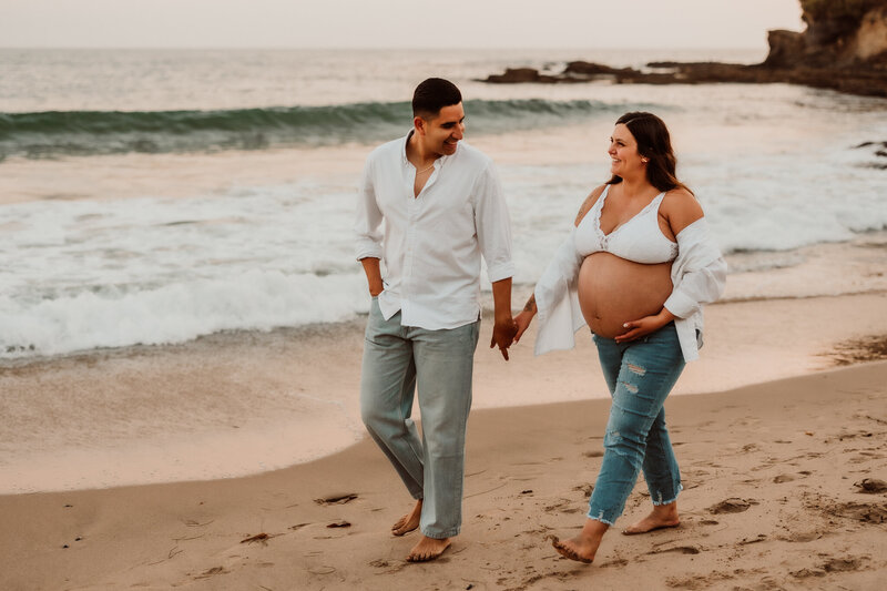 Pregnant couple walking barefoot on Laguna Beach during a sunset maternity photo session.

