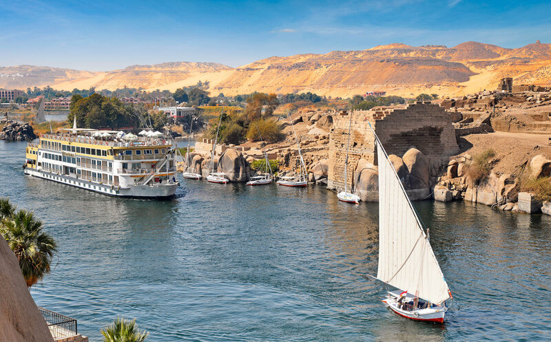 A scenic view of the Nile River with a large cruise ship and a traditional sailboat (felucca) gliding through calm blue water, surrounded by rocky shores, palm trees, and golden desert hills in the background.