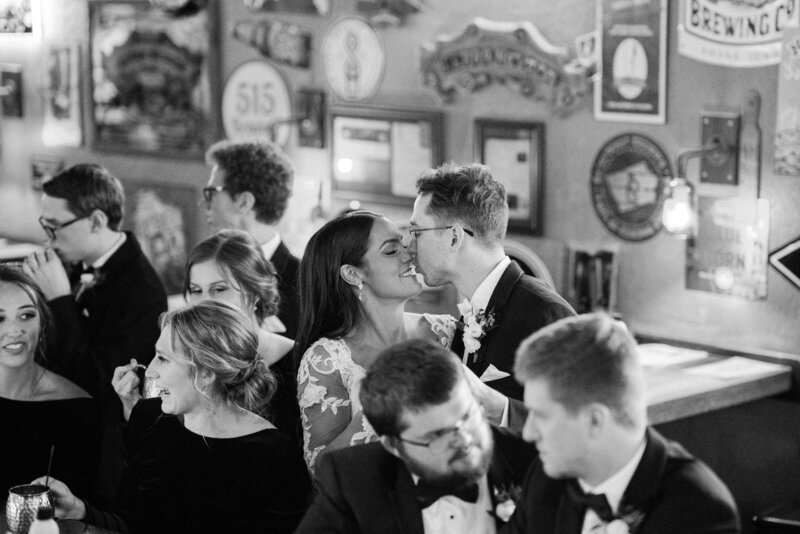 A couple enjoy a quick kiss as they hang out in a bar with all of their friends after their wedding ceremony
