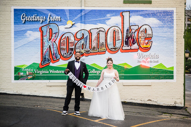 An elopement couple stand in front of a mural in Downtown Roanoke that says "Greetings from Roanoke: Star City of the South." The couple is smiling and holding a sign that says "just married."