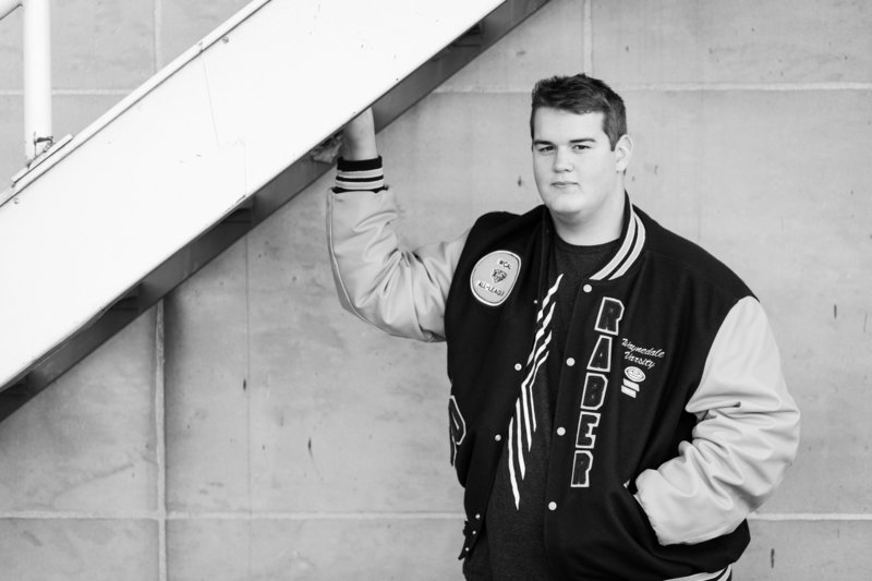 black and white photo of senior guy wearing letterman jacket in front of stone wall, photographed downtown Wooster, photographed by Jamie Lynette Photography Canton Ohio Senior Photographer