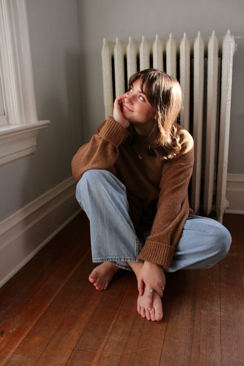woman wearing a brown sweater and jeans sitting on the floor in front of a radiator with her chin in her hand