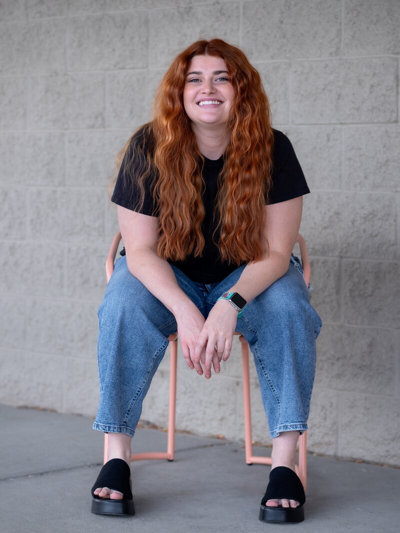 branding headshot portrait of a hairstylist. she has long red curly hair and sits on a chair, leaning forward on her elbows.