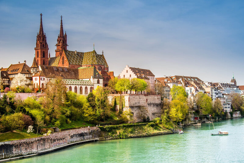A riverside view of a historic European city featuring a large cathedral with twin spires, traditional buildings, lush trees, and calm turquoise water under a clear sky.