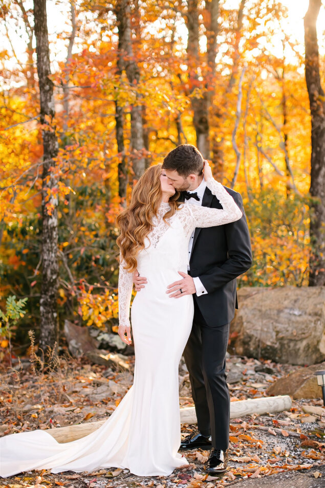 A bride and groom kissing with trees behind them during Fall.