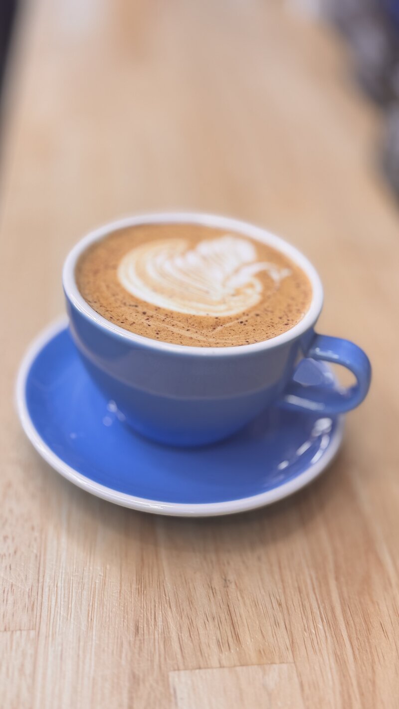 A cup of cappuccino with intricate latte art, topped with mini marshmallows, sits in a cozy coffee shop in Temple, Texas. Surrounded by coffee beans on a dark surface, the scene is accented by a burlap cloth and autumn leaves fading into the blurred background.