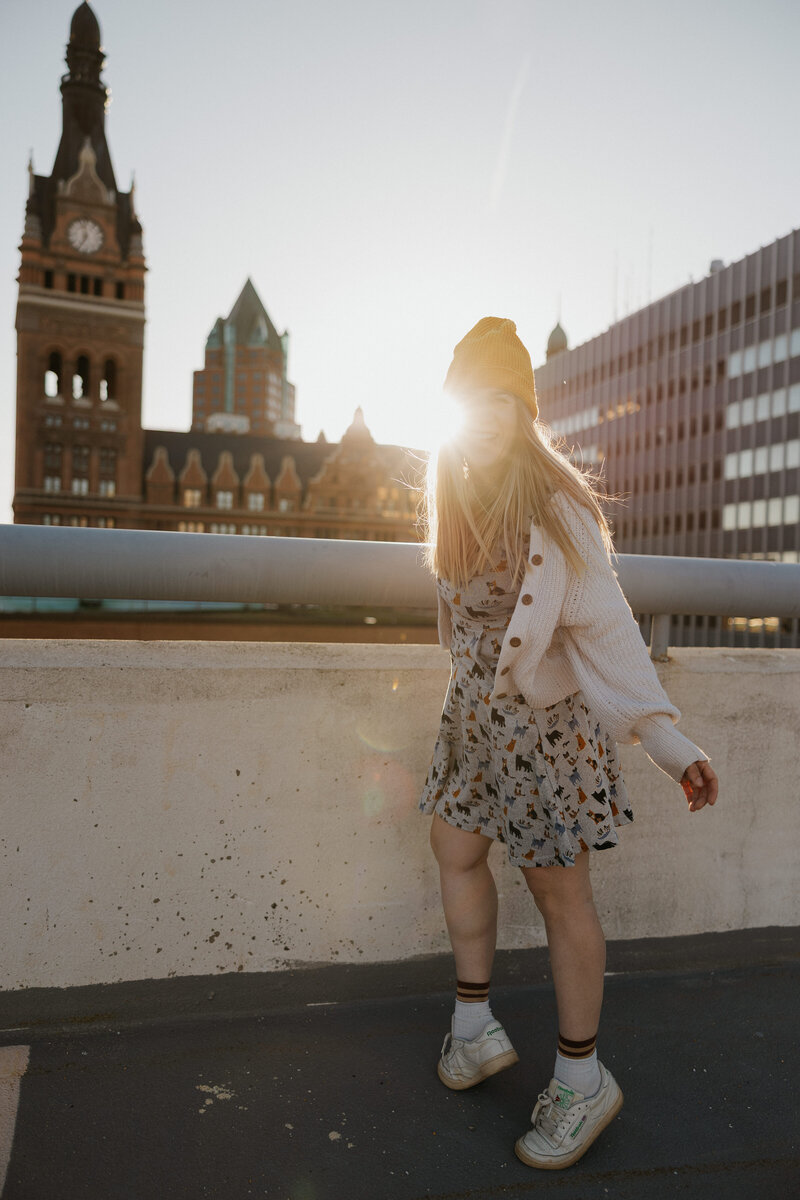 A photographer holding up a camera and smiling as they ride a skateboard 