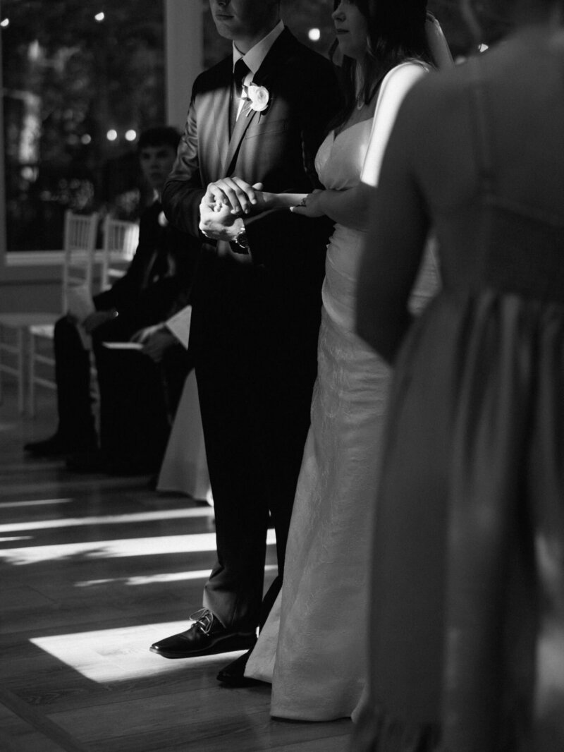 Intimate black-and-white photo of a bride and groom holding hands during the ceremony.