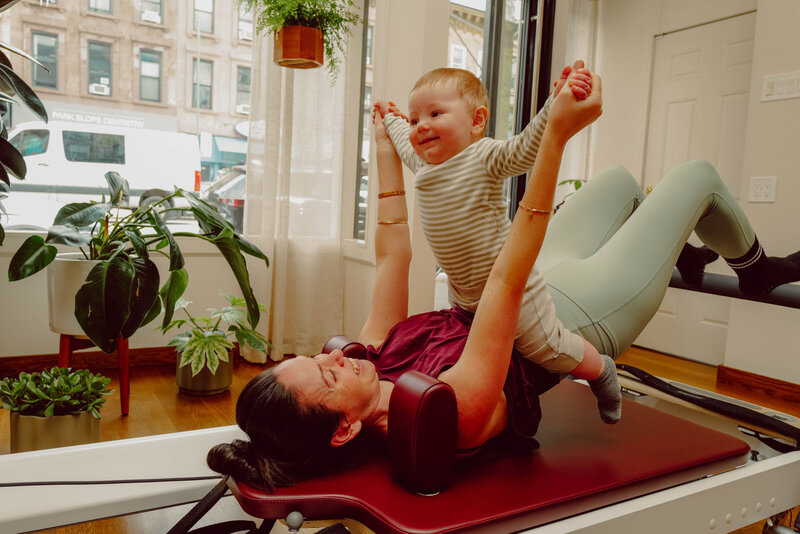 A mom holding her baby while laying on the reformer to a Baby and Me Pilates class in Park Slope, Brooklyn