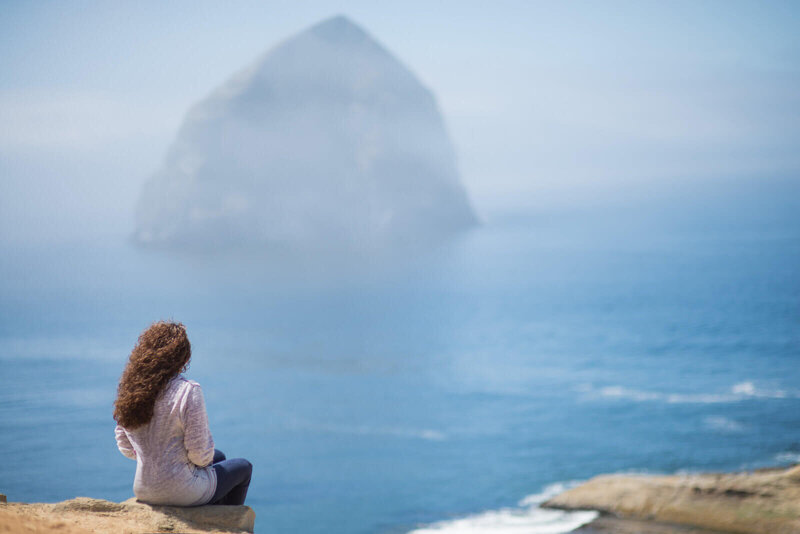 A person with long curly hair and a light-colored shirt sits on a rocky ledge overlooking a foggy ocean. In the distance, a large, mist-shrouded rock formation rises from the water, creating a tranquil, contemplative scene.