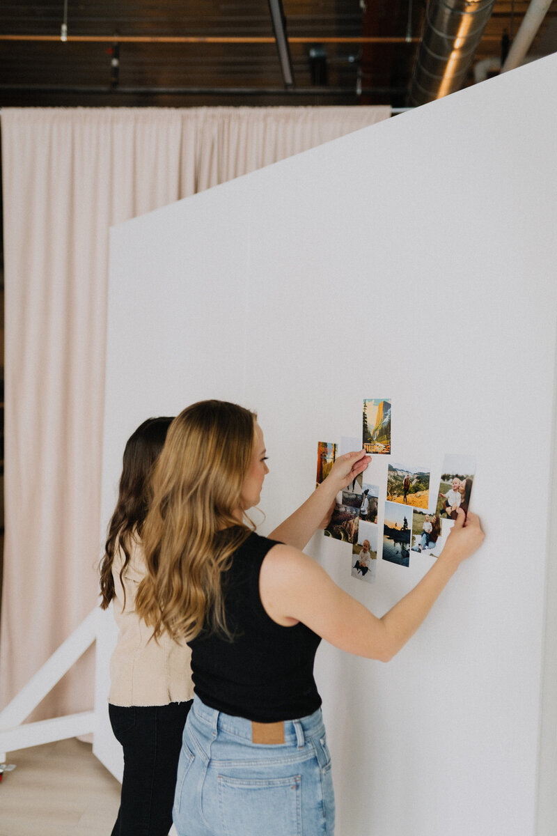 Two women pasting travel postcards and photos onto a white wall