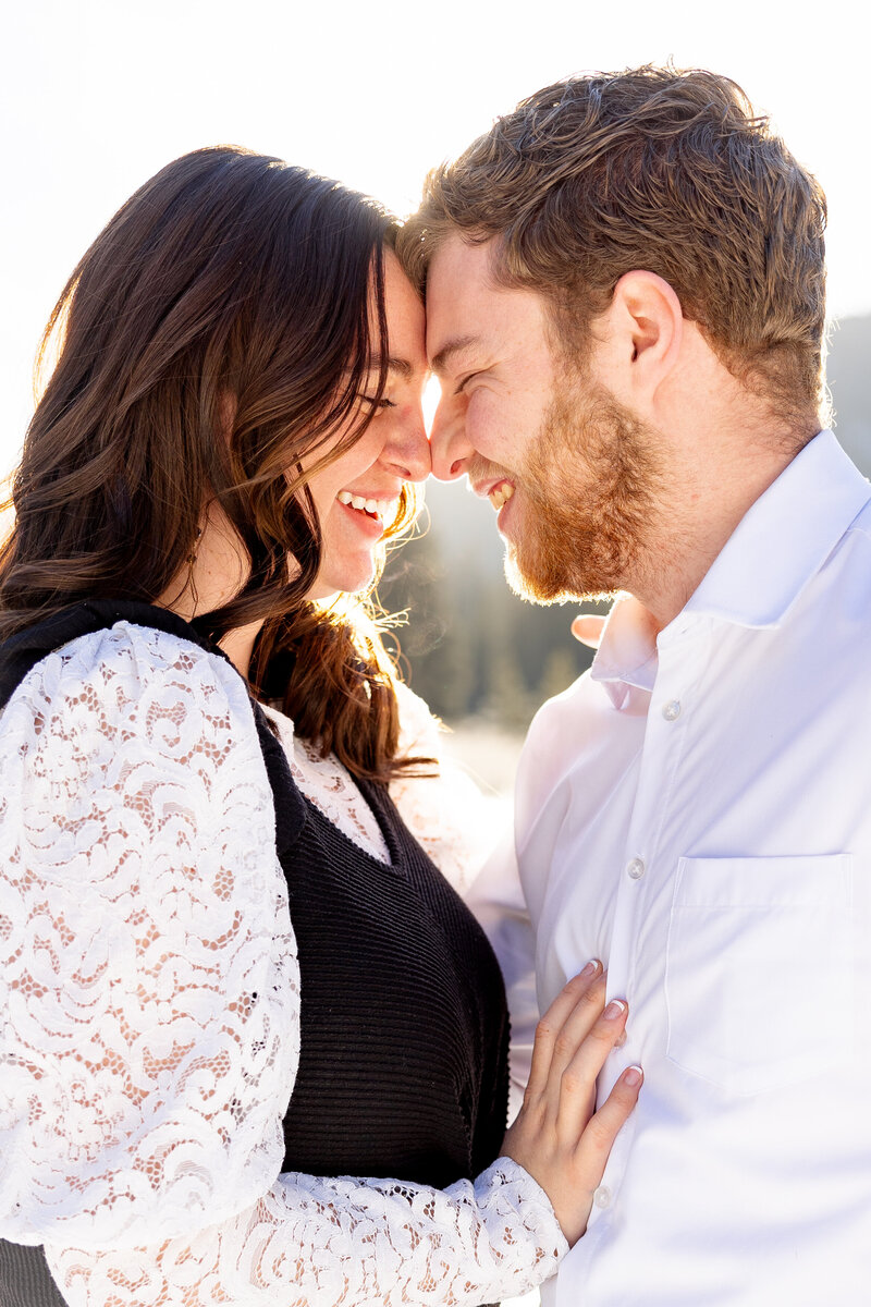 The engaged couple touches noses and smiles at each other in the beauitful sunlight up in the snowy mountains.