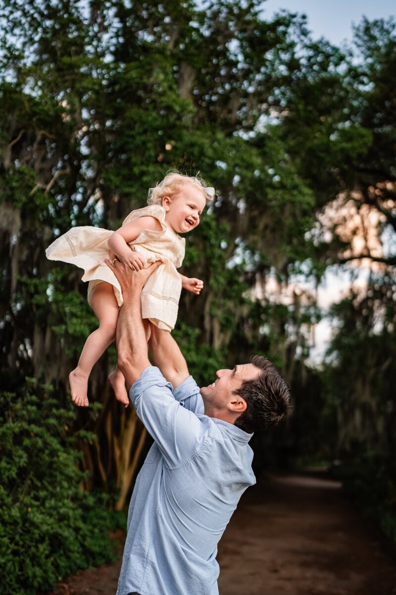 Dad lifting toddler daughter joyfully into the air under mossy oak trees during a golden-hour family photo session in Charleston.