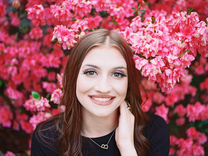 Girl smiling in front of bright pink flowers and hand on her neck
