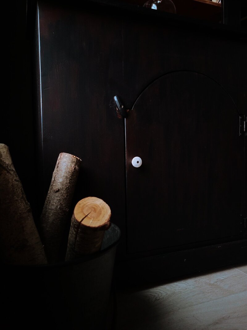 Black chunky cabinet with arched door and white floor with a tin filled with white birch logs