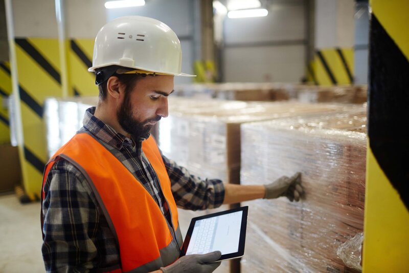 A man in a hard hat is using a tablet in a warehouse.