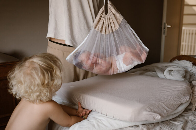 Baby wordt gewogen in een doorzichtige doek. Grote broer kijkt toe.