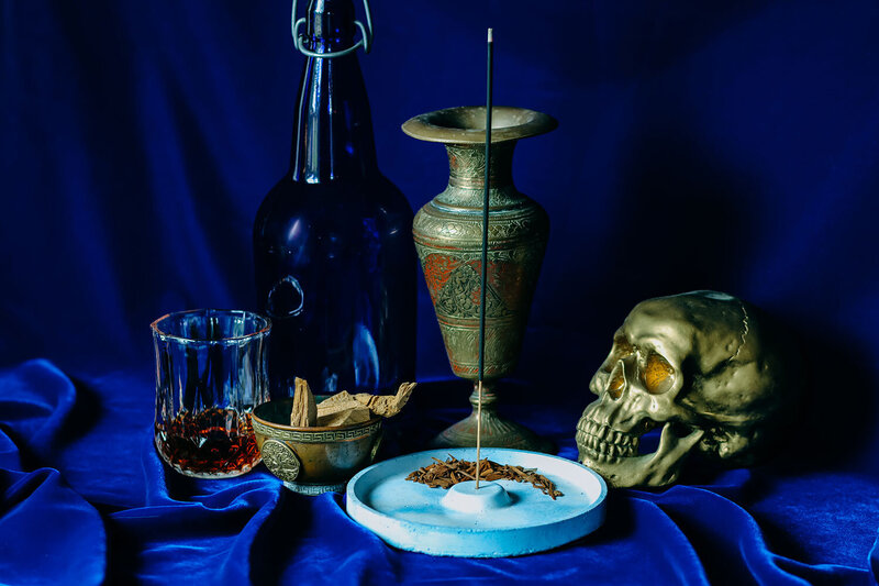 A still life centering a single, lit stick of charcoal incense. The background is a deep blue velvet. Around the incense in its burner is sticks of oud and cedar in an ornate brass offering bowl, a dark blue old swing-top bottle, a faded brass vase, a golden skull, and whiskey in a neat glass.