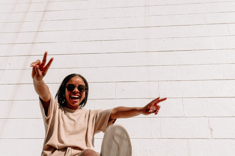 A black woman smiles as she poses against a white brick wall, she's wearing a tan shirt and is doing the peace sign with both hands