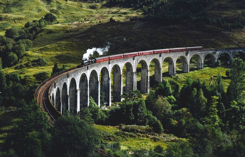 A vintage steam train glides across the iconic Glenfinnan Viaduct, surrounded by lush green hills in the Scottish Highlands. 