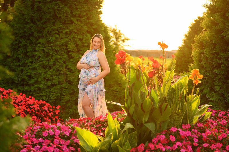 Blonde woman cradling her belly with the sun rising behind her and flowers and trees all around for her outdoor Burlington, On Maternity Session.