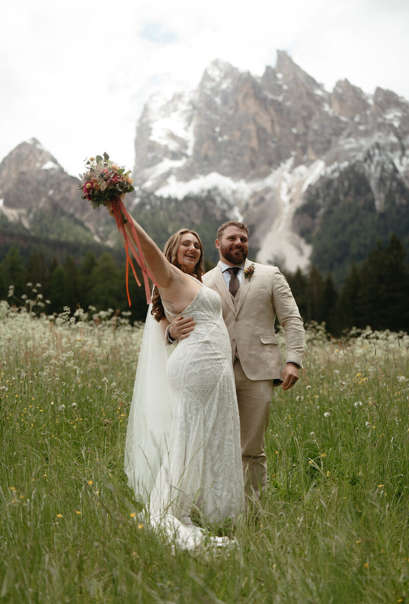 Bride holds her bouquet from a Dolomites Florist up above her head with a huge smile on her face 