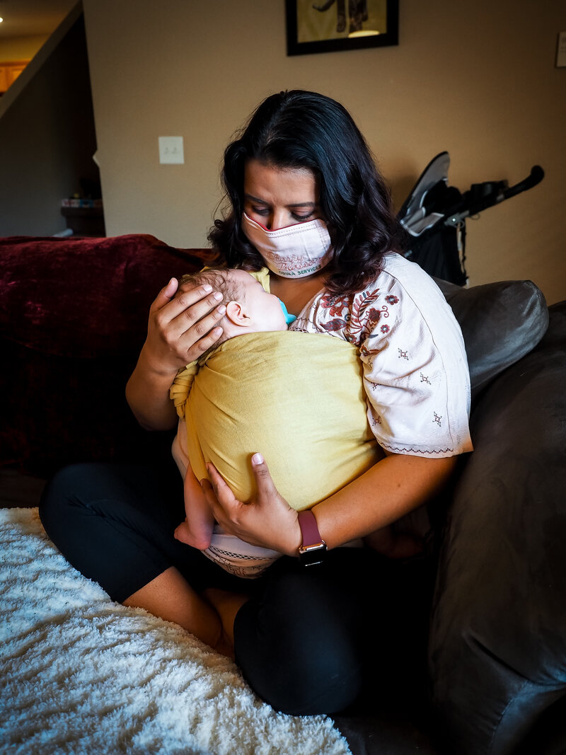 Woman wearing mask holding baby in a yellow wrap