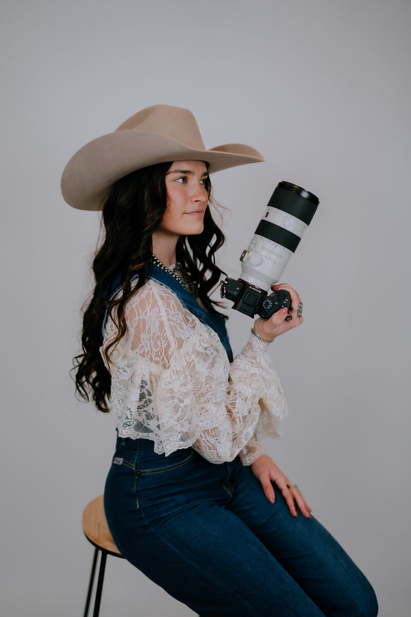woman holding a camera while sitting on a stool wearing a cowgirl hat