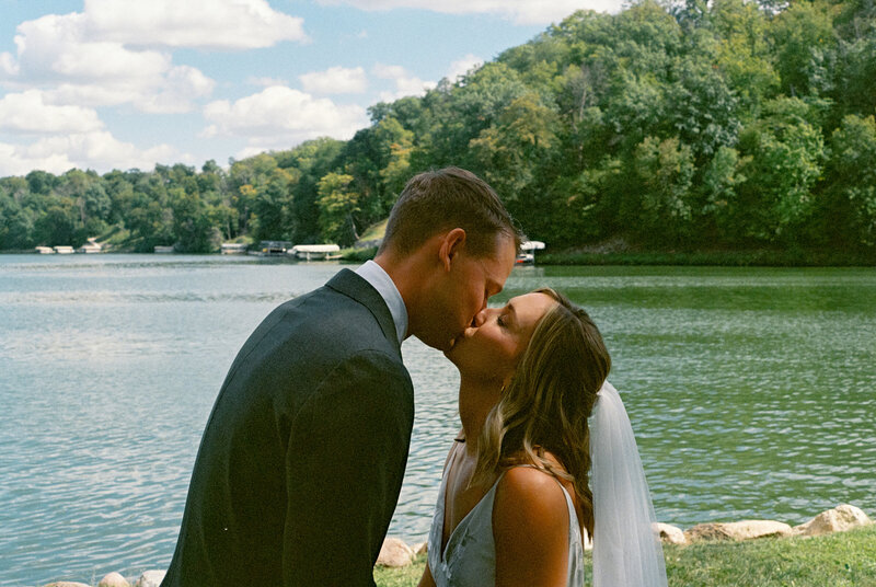 Bride and groom share a kiss by a peaceful Iowa lake, surrounded by green trees and under a bright blue sky.