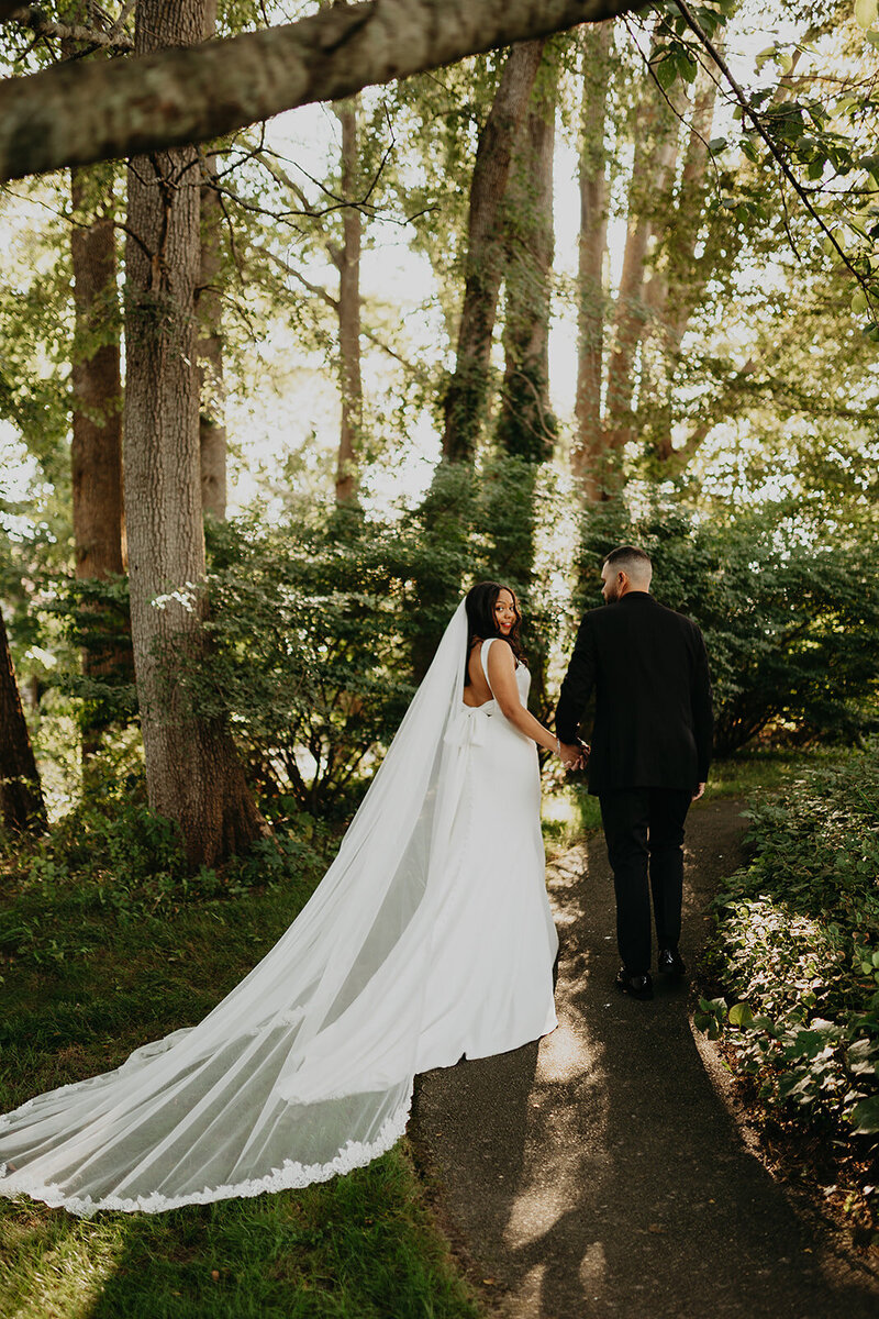 At the Coonamessett Inn wedding venue, the newlyweds stroll through the forest, hand in hand. The bride turns with a smile, capturing a moment of pure joy.
