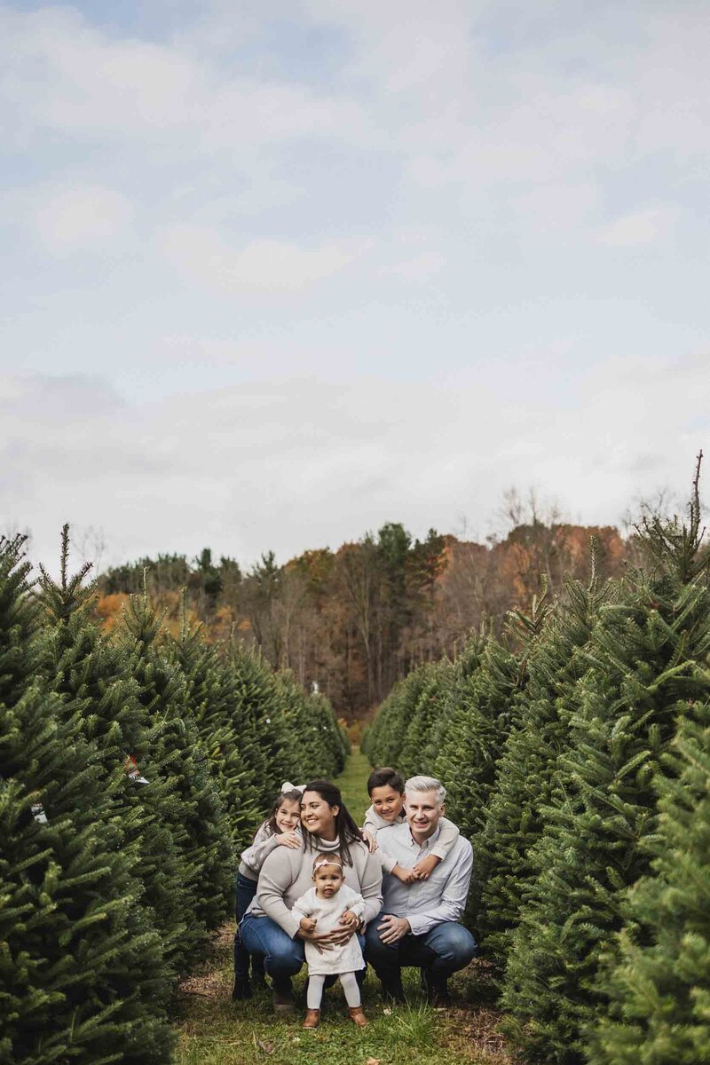 Family of five crouching together between rows of evergreen trees during a Christmas mini session at a tree farm in Northeast Ohio, with autumn foliage in the background.