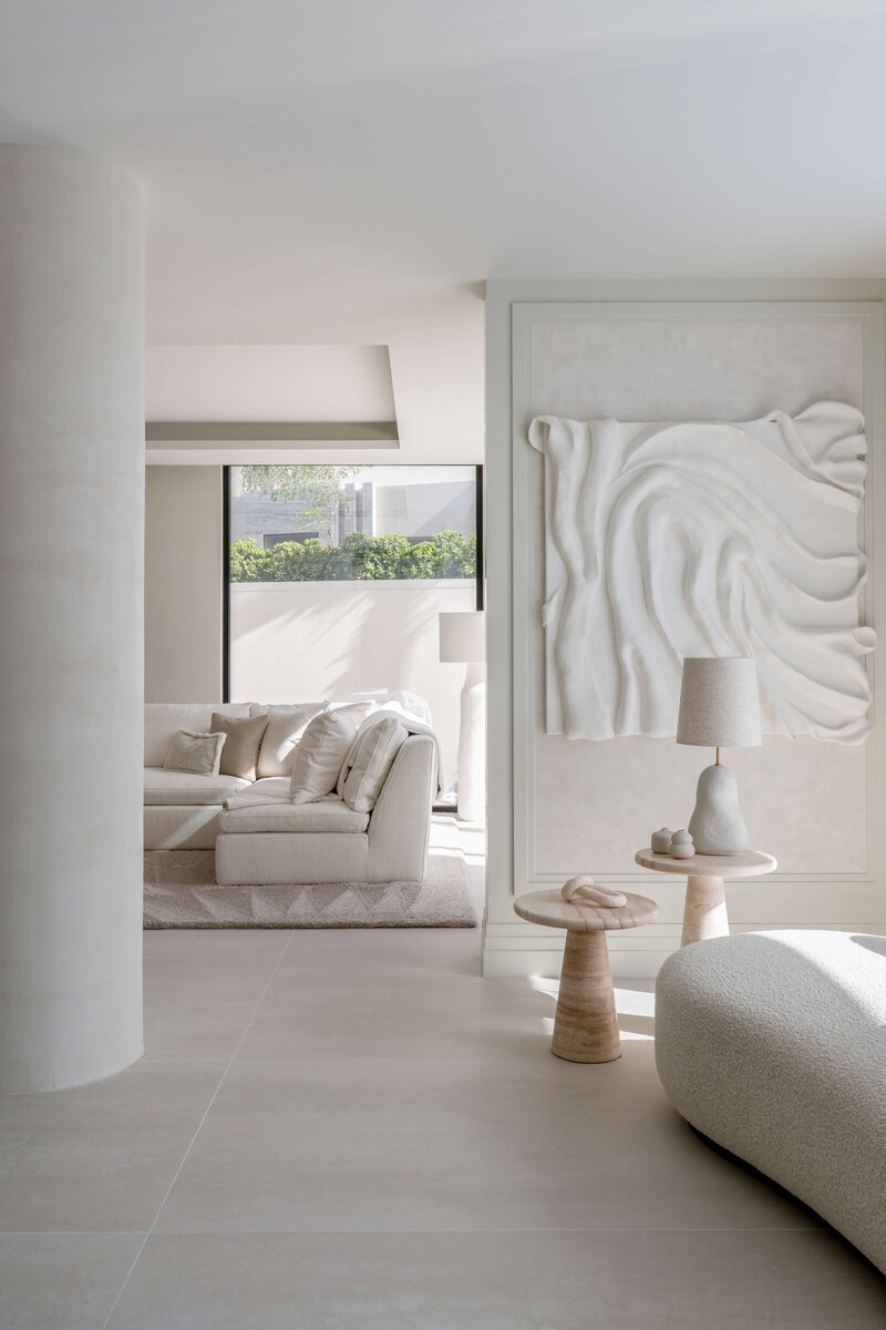 A white wall with a textured white art piece. Beyond, a glimpse into the light, airy living room featuring a white couch and light colored rug.