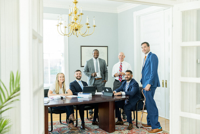 Professional law firm team gathered in a bright, elegant office space in Columbia, SC, featuring a gold chandelier, classic white trim, and a polished wooden conference table. Attorneys in suits and business attire present a confident, approachable image. Small business branding photography in Columbia, SC, specializing in law firm branding and corporate professional imagery.