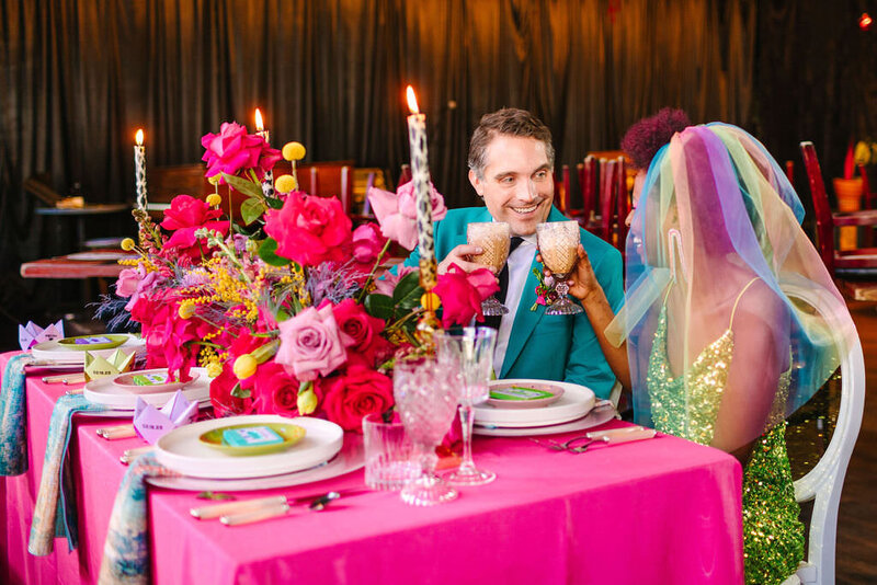 A wedding couple sitting at a reception table holding up glasses.