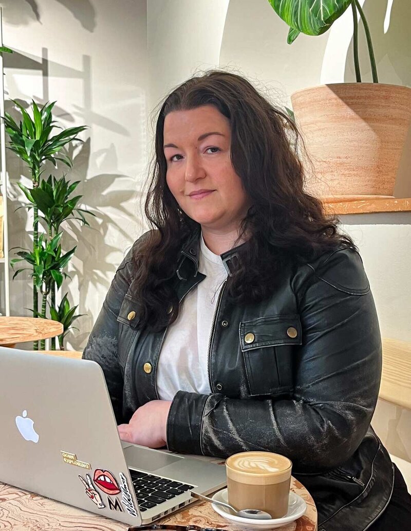 Martina Menzini wearing leather jacket, writing and consulting on laptop in cafe, holding a latte and surrounded by plants.