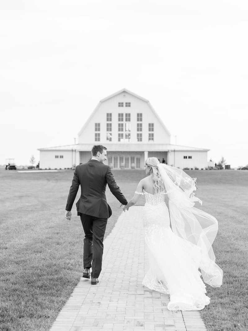 Bride and groom run through a field toward a white barn wedding venue