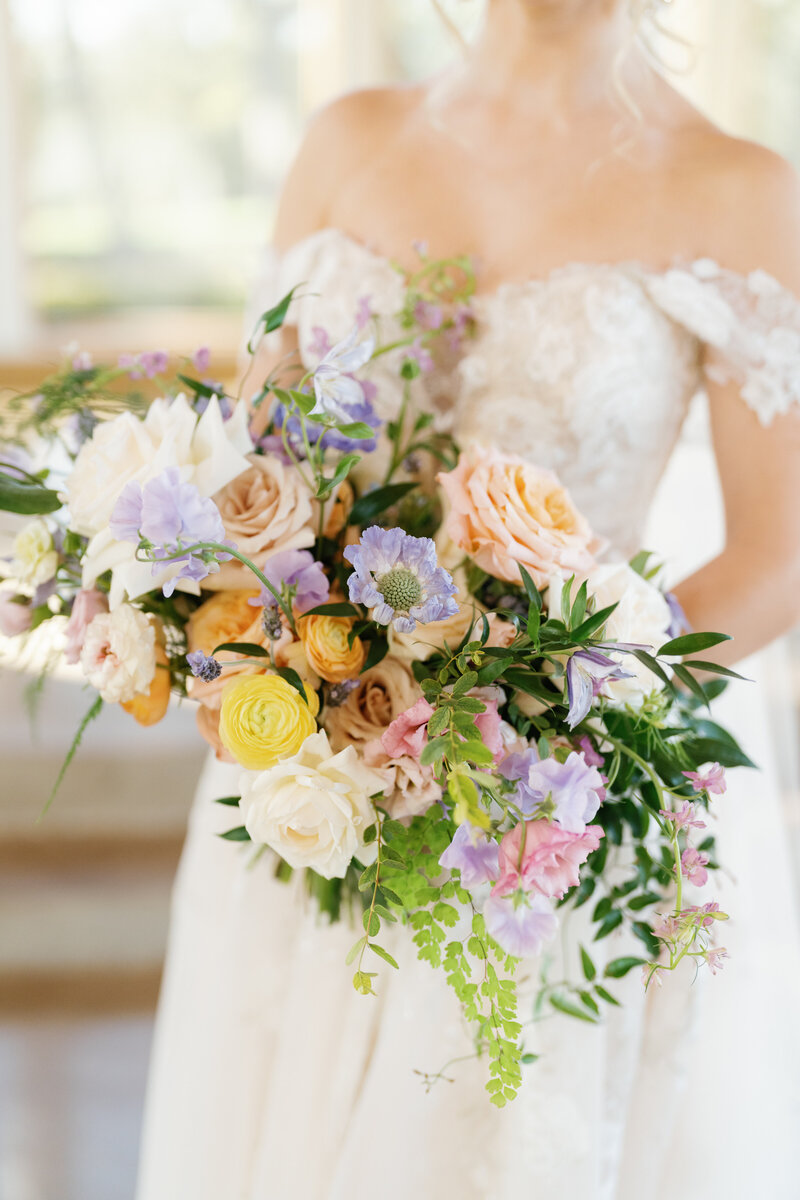 Close up image of a bride holding a colorful bouquet of orange, purple, and white flowers with greenery