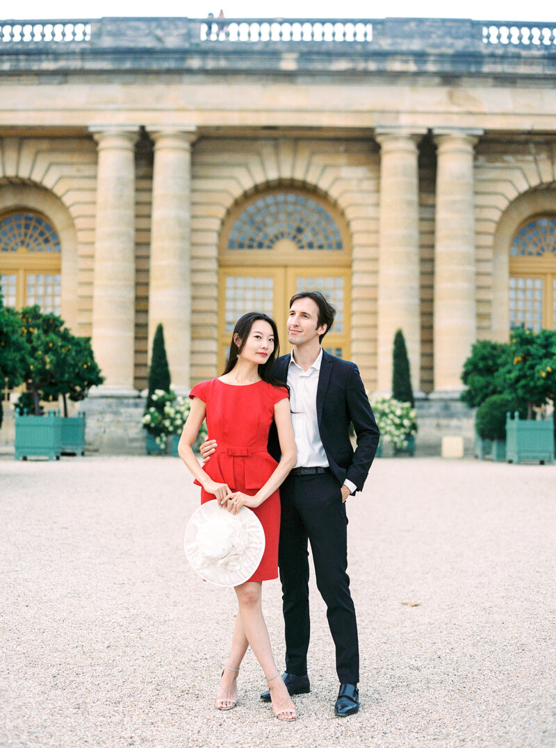 The Future Bride and Groom in the château de Versailles for the engagement photoshoot, red dress and black suit, near Airelles Versailles, Orangerie garden