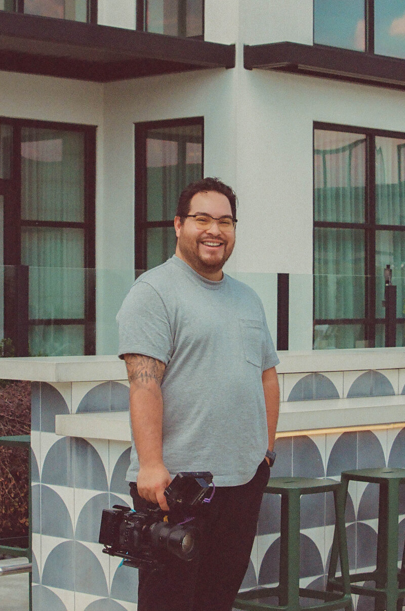 A man stands with a camera in front of a house, smiling