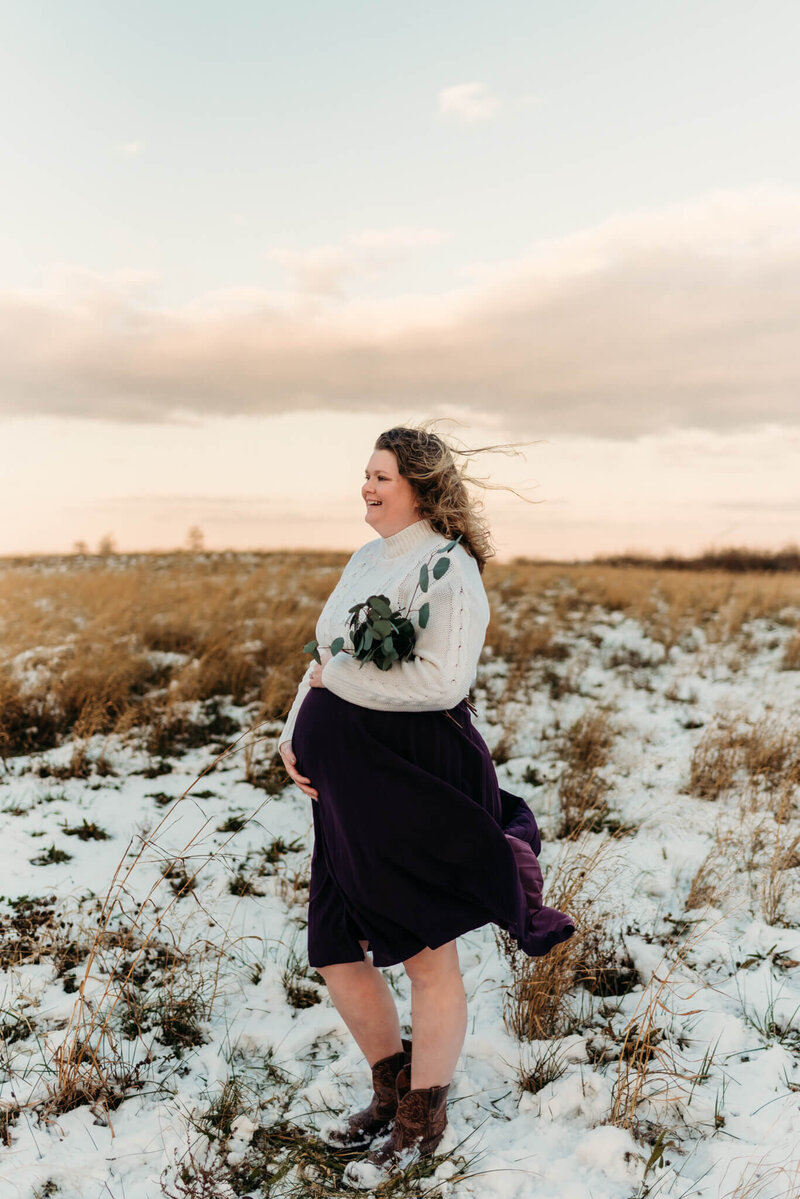 Pregnant mom standing in a field with a light layer of snow.  She is cupping the under side of her baby bump while the wind blows her hair and purple skirt.  Photo take by Delaware maternity photographer, Kristi