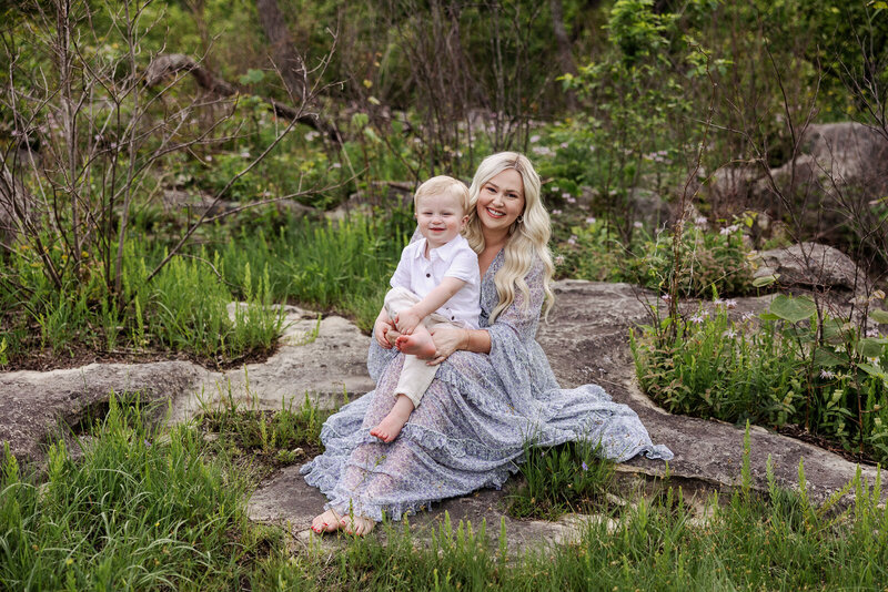 Portrait taken of mother and toddler sun in the spring sitting on rocks surrounded by green grass and flowers in De Soto, Missouri. 