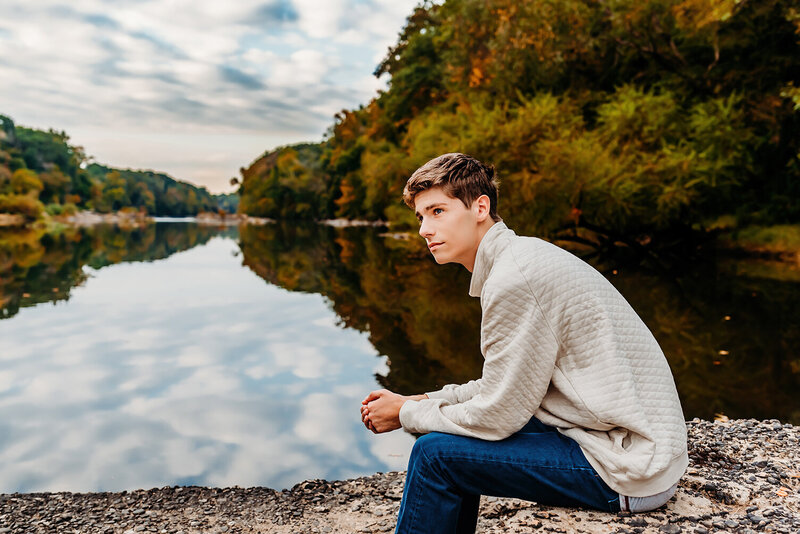 a senior boy sitting on a rock on a river in the fall