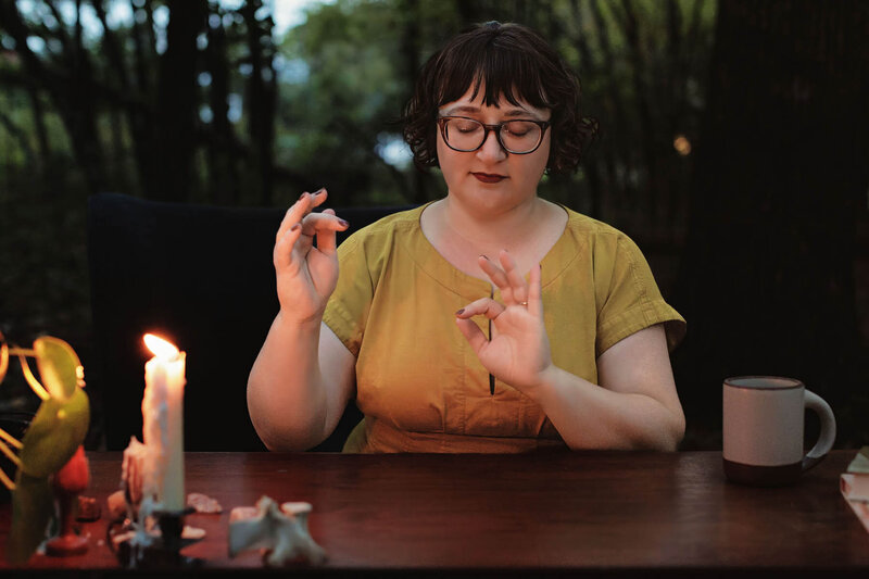 Ash sits at a wood table in the trees practicing energy work wearing a mustard yellow dress