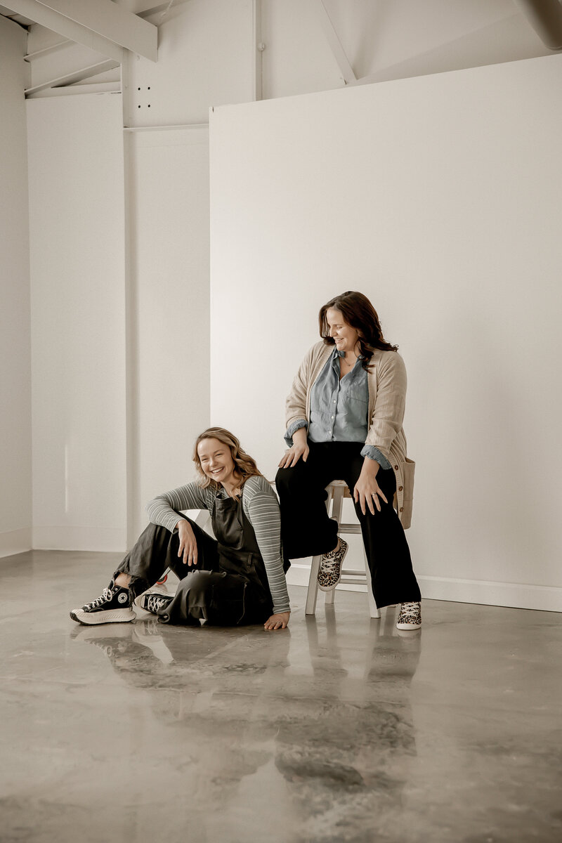 Lacey sitting on a stool looking down at Amber sitting on the floor, laughing together