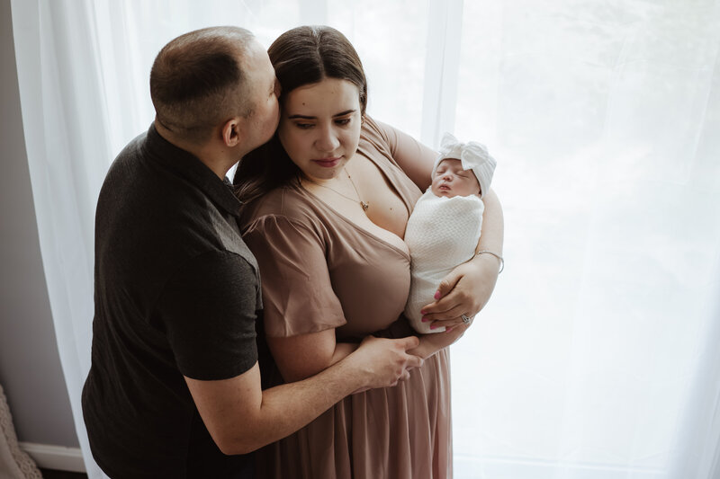 Dad is black shirt kissing the temple of mom wearing a pink dress while holding baby wrapped in white blanket next to a window.