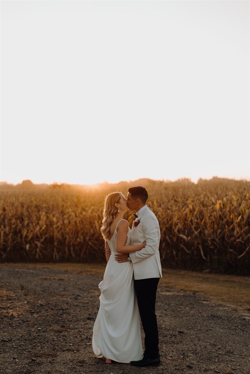 A bride and groom wearing a white dress and white suit standing together at sunset next to a corn field at The Narrows Landing a prominent Hamilton Wedding Venue